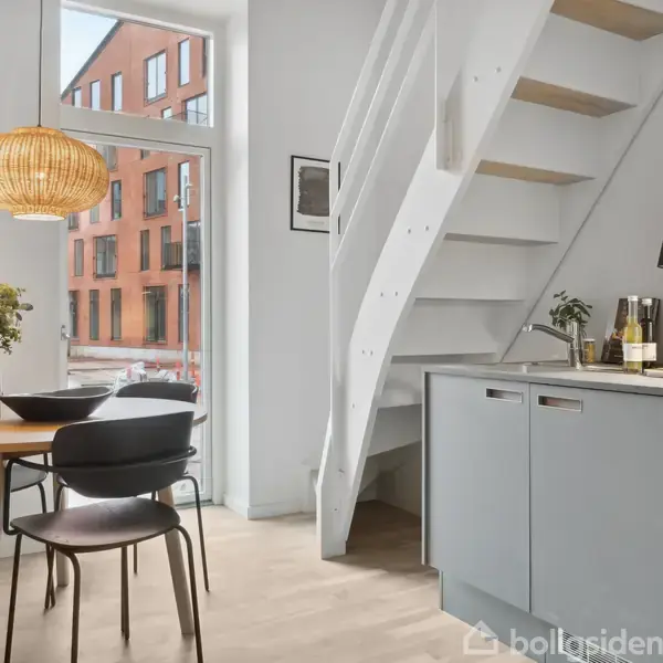 Et moderne køkken med grå skabe og træbord. To stole står ved bordet, dekoreret med planter. En trappe leder opad. Udenfor ses en murstensbygning gennem vinduet.