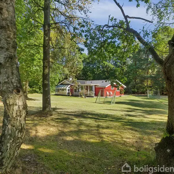 Et sommerhus med et drivhus-lignende tilbygning står i en solrig have omkranset af høje træer. Verandaen vender mod det åbne græsareal og skovklædt baggrund.