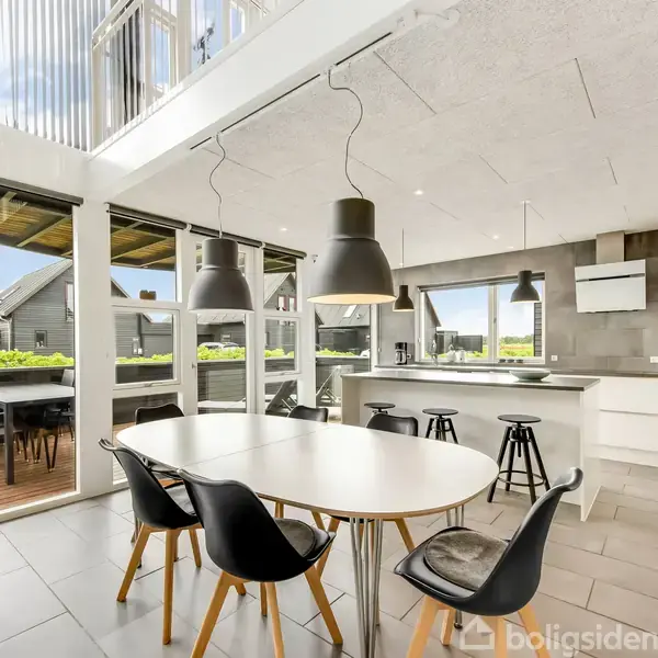 Et moderne køkken med en oval spisebord omgivet af sorte stole. Store vinduer tillader udsigt til en terrasse med siddepladser. Lysegrå fliser og indbygget køkkenudstyr skaber et stilrent miljø.