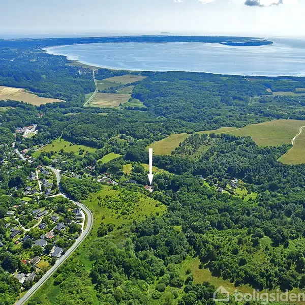 Et landskabsbillede viser grønne skove og marker med en vej, der snor sig gennem en landsby til venstre. En pil peger mod en specifik placering, mens en bugt og ø er synlige i baggrunden.
