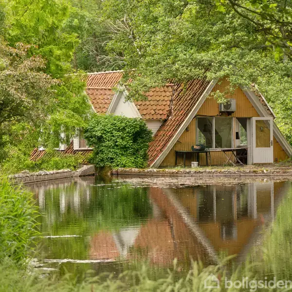 En A-formet hytte med store vinduer står ved en stille sø. Hytten er omgivet af tæt, grøn skov, og dens tag er dækket af røde teglsten.