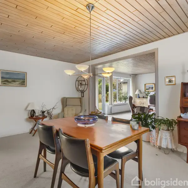 Et træbord med to stole står i en spisestue. Ovenfor hænger en fem-armet loftslampe. Rummet har en moderne stil med hvide vægge, trægulve og forskellig dekoration.