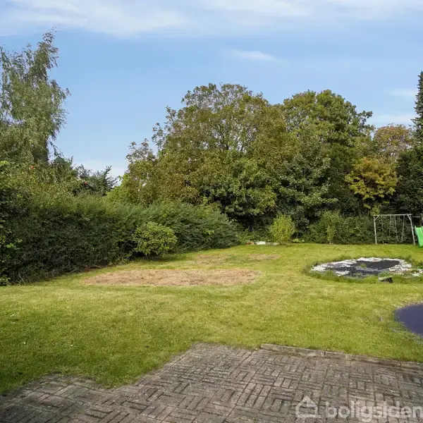 Græsplæne strækker sig fra terrasse med fliser til en cirkulær bålplads, omkranset af træer og buske; en gynge hænger i baggrunden.