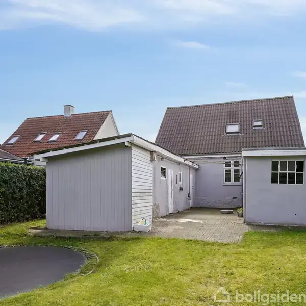 Et hvidt hus med brun tag udfører ingen aktivitet. Det omgives af en velholdt græsplæne med en trampolin i venstre side under en blå himmel.