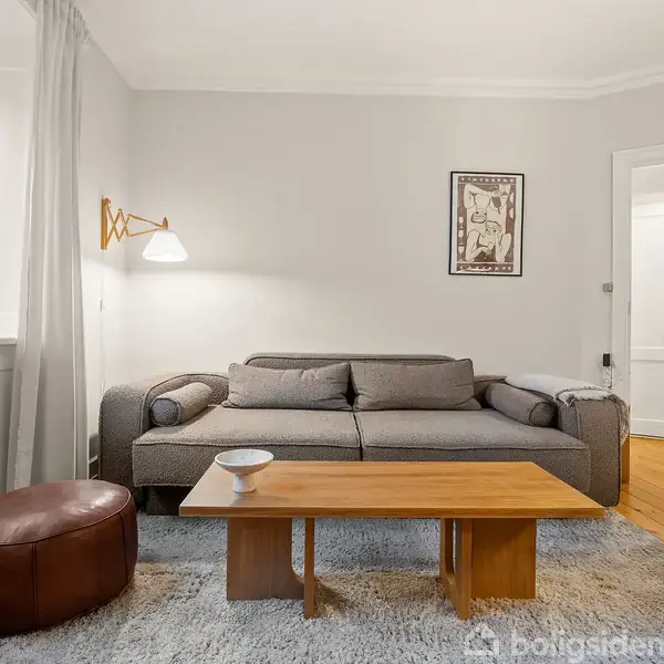 En grå sofa står i en stue ved siden af et brunt læderpuf. Et træbord med en skål står foran sofaen. Væggene er lyse, og et billede hænger ovenover sofaen. En lampe lyser fra venstre. Et "Til salg" skilt ses gennem vinduet.