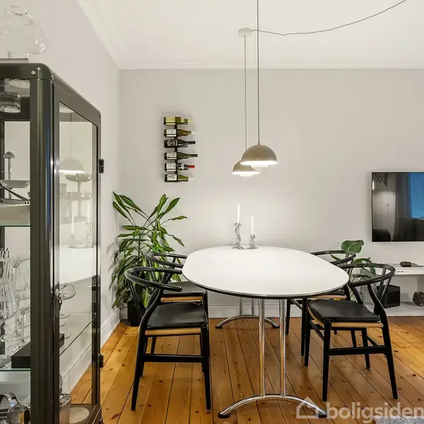 En spisestue med et rundt hvidt bord omgivet af sorte stole. En glasvitrine viser glasservice. Der hænger en lampe over bordet. En væg har et væghængt TV og en vinholder.
