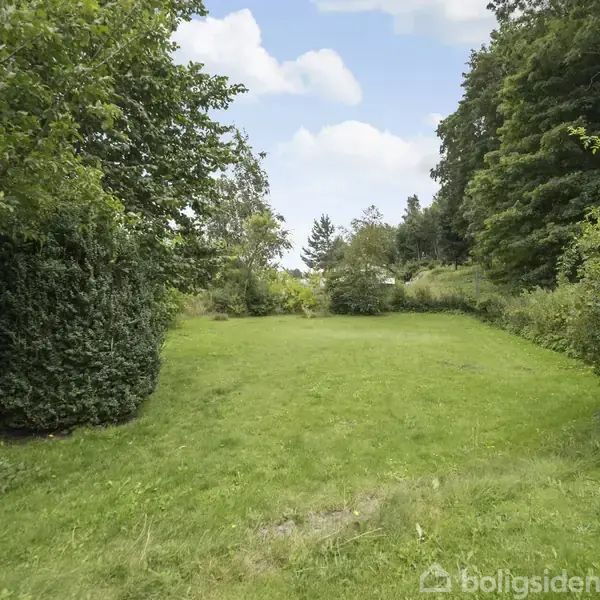 En græsplæne strækker sig midt i en have, omgivet af tæt, grøn beplantning og træer. Himlen er klar med enkelte skyer, der skaber en fredfyldt atmosfære.