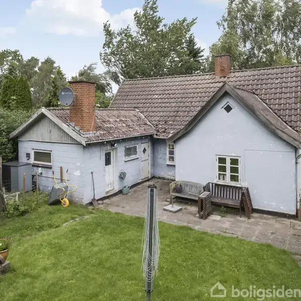 Et lille hvidt hus med tegltag står på en græsplæne omgivet af træer. På terrassen er der en bænk og en parasolfod.