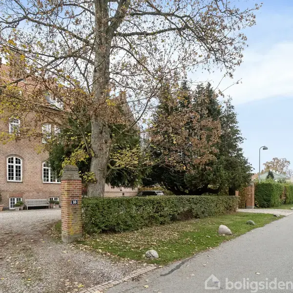 Stort rødt murstenshus med karnap og spidst tag i stille, kystnært område med udsigt til havet. Omgivet af træer og lille have, nær marina.