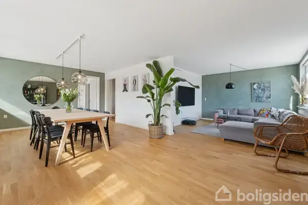 Et lyst, åbent opholdsrum med spisebord og stole til venstre. En stor plante står i midten. Til højre ses en hjørnesofa og moderne indretning.
