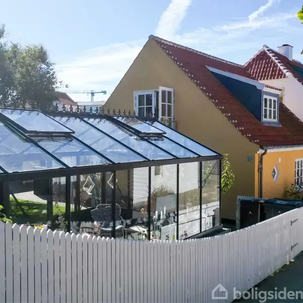 Et gult hus med rødt tag står ved siden af et drivhus i glas, omgivet af et hvidt hegn, i et solrigt boligområde med træer og blå himmel.