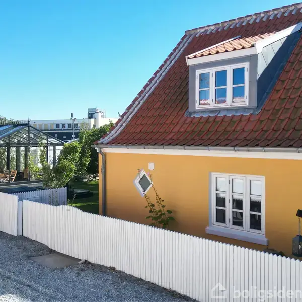 Gul hus med rødt tegltag står stille ved en hvid stakit i en gade. Bagved ses et drivhus og grøn have under en klar blå himmel.