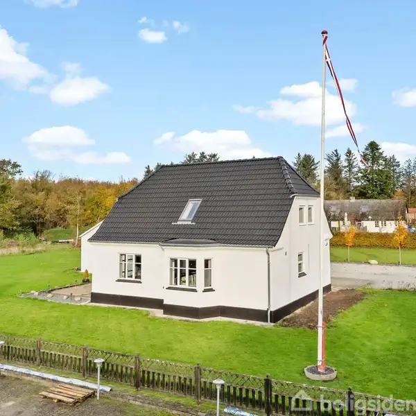 En hvid, to-etagers bolig med sort tag står på en velplejet græsplæne. En høj flagstang med flag er i nærheden, omgivet af skovbevoksning og en blå himmel med skyer.