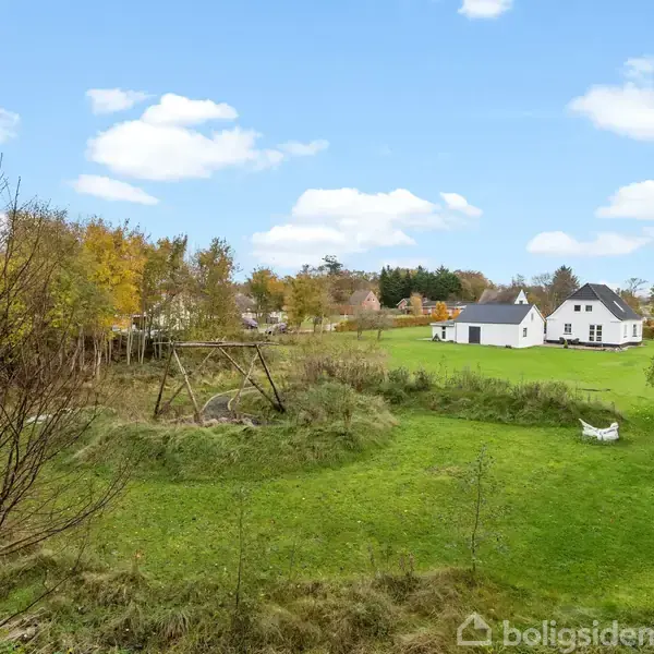 En stor græsplæne med en tom metalgynge i midten, omgivet af træer. I baggrunden ses et hvidt hus med et mørkt tag, under en blå himmel med skyer.