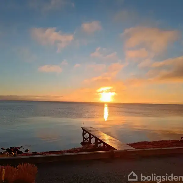 En træbro strækker sig ud i en stille sø, mens solen går ned over horisonten og skaber en gylden refleksion. Skyer svæver i en lyserød-orange himmel.