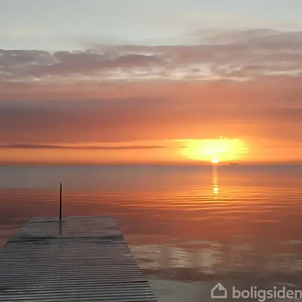 En træbro strækker sig ud mod en stille havoverflade ved solnedgang, med en orangerød himmel og solen, der lige er ved horisonten. En båd ses i det fjerne.