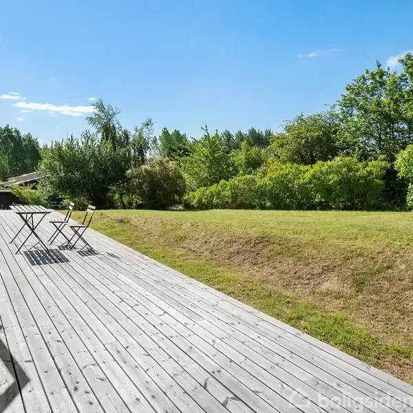Terrassen rummer to stole og et bord i foldbare stil, stående på trædæk, omkranset af en veltrimmet græsplæne og omgivet af frodige buske under en klar blå himmel.
