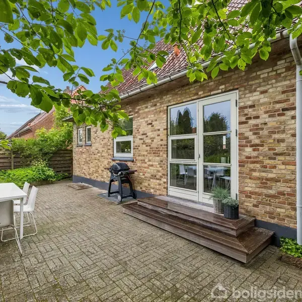 Hus med grå facade og rødt tegltag står foran en velholdt have med buske og træer. I højre side er en carport med biler ved siden af.