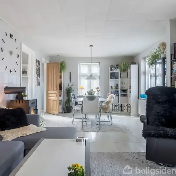 Stue med grå sofa og lænestol ved et sofabord. I baggrunden ses en spisestue med et bord og stole. Hylder med bøger og planter omkranser rummet.