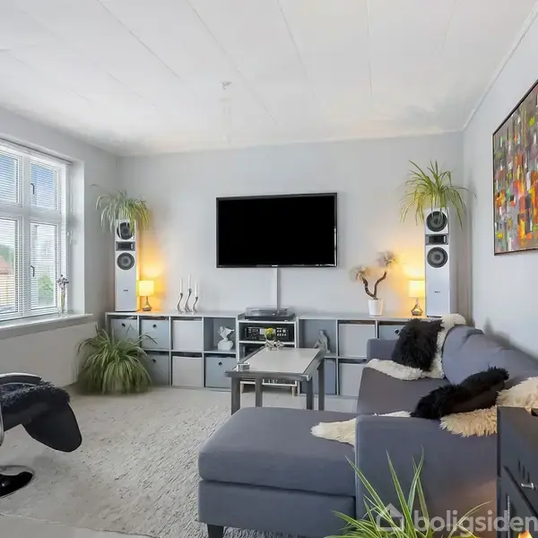 Stue med en grå sofa i hjørnet, fladskærms-TV på væggen, planter over reoler, sort lænestol ved vindue, og farverigt maleri. Moderne og lys atmosfære.