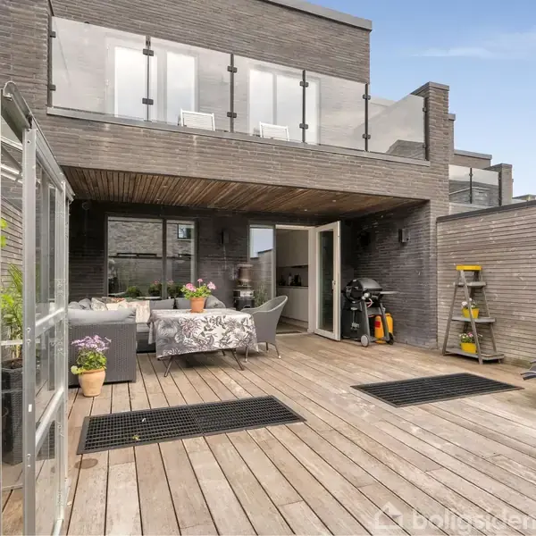 En træterrasse med havemøbler og planter, omgivet af moderne murstensbygning med altan. Et lille drivhus til venstre og redskaber op ad væggen til højre.