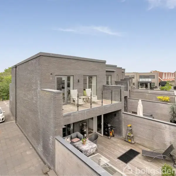 Moderne murstenshus med to terrasser, den ene med møbler, i et boligområde. Foran huset er en flisebelagt indkørsel med en parkeret bil. Grønt område i baggrunden.