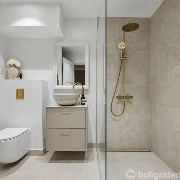 Et moderne badeværelse med en væghængt toilet, en vask på et skab, og en bruseniche med gyldne armaturer. Rummet har lyse fliser og minimalistisk indretning med pæne detaljer.