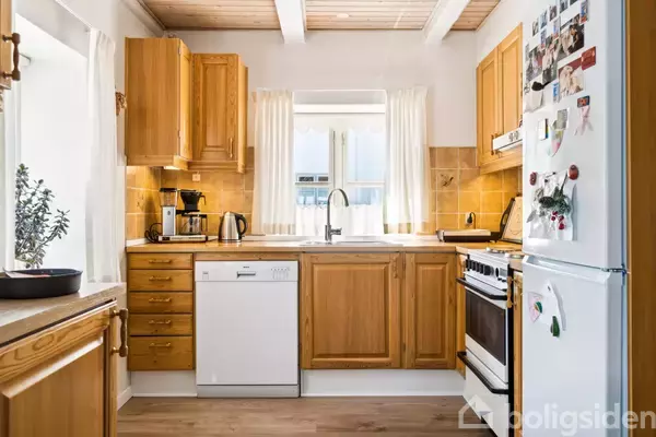 Køkken med træskabe, opvaskemaskine, og hvidevarer står stille i et lyst rum. Vinduet med hvide gardiner lader naturligt lys ind. Køleskabet har magneter og billeder.