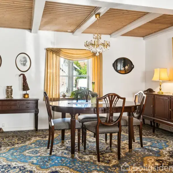 Et spisebord med stole står midt i et rum med tæppe. Væggene er dekoreret med malerier og figurer. En lysekrone hænger over bordet, og en kommode med lamper står til højre.