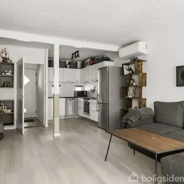 Grå sofa står i moderne stue, ved siden af træbord. Køkken med hvide skabe og stål køleskab i baggrunden. Hylder med dekorationer fylder væggen. Mørkt trægulv.