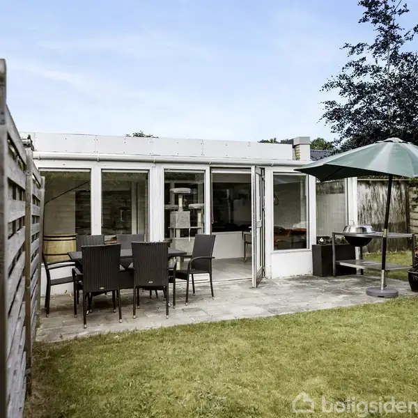 Terrassemøbler står på flisebelagt område foran et sommerhus med store vinduer. En parasol og grill er placeret tæt ved. Græsplænen omgiver terrassen, og der er hegn i baggrunden.