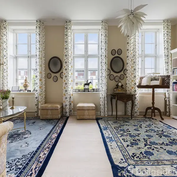 Et elegant stuearrangement med sofa, sofabord og blå tæpper. Tre store vinduer med mønstrede gardiner lader lys ind. Hylder med bøger og vægdekorationer skaber en hyggelig atmosfære.