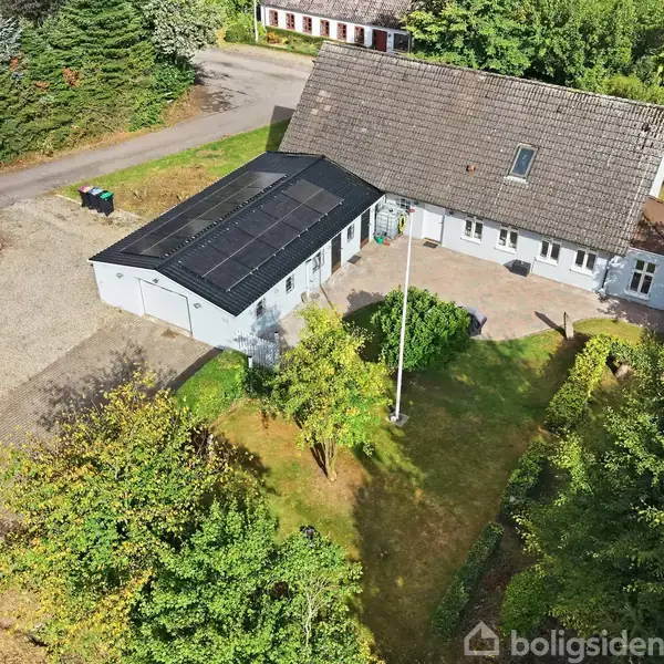 Hus med solpaneler på taget set fra luften, omgivet af en have og træer. Grusindkørsel og vej i nærheden, giver et landligt indtryk.
