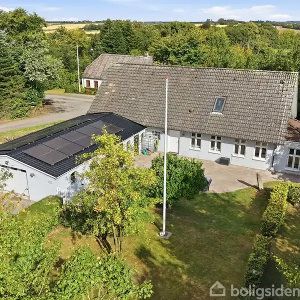 Hus med solpaneler på taget set fra luften, omgivet af grønne træer og buske. Huset ligger på en velplejet græsplæne med flagstang, i en landlig, skovklædt område.