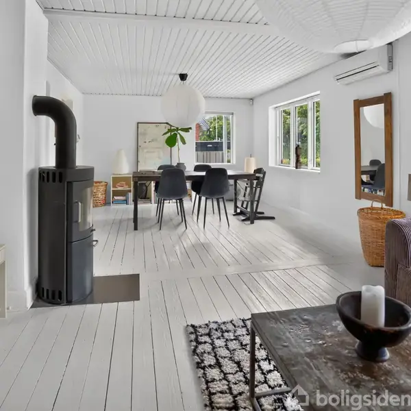 Stue med hvidmalede trægulve og vægge indeholder sort brændeovn, spisebord med sorte stole, stor papirlampe, lilla sofa og bord. Vinduerne giver naturligt lys.