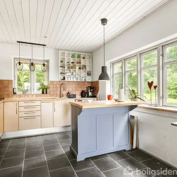Et lyst køkken med beige skabe og en blå køkkenø. Vinduer giver udsigt til en grøn have. Der er mørke fliser på gulvet og moderne lamper over arbejdsområdet.