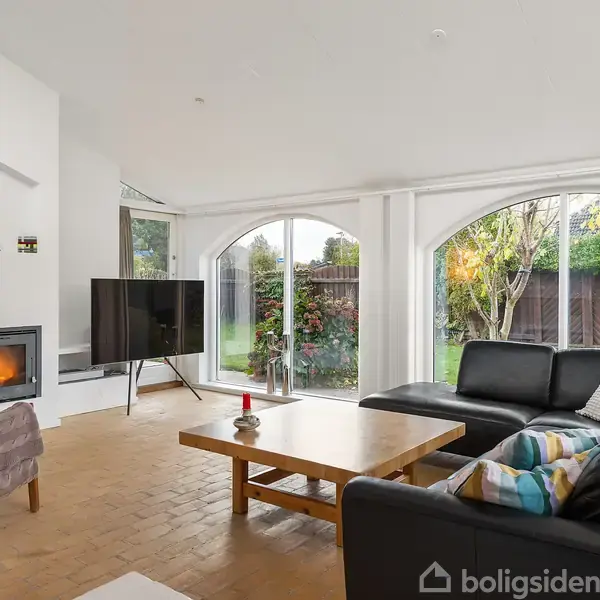 Brændeovn oplyser stue med sorte lædersofaer og træbord, mens stort vinduesparti viser have med buske og træer. Fjernsyn står til venstre ved siden af pejsen.