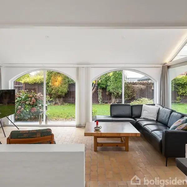 Stuen indeholder en sort lædersofa og et sofabord foran store vinduer mod en grøn have. En pejs er til venstre, og et tv står på et stativ.