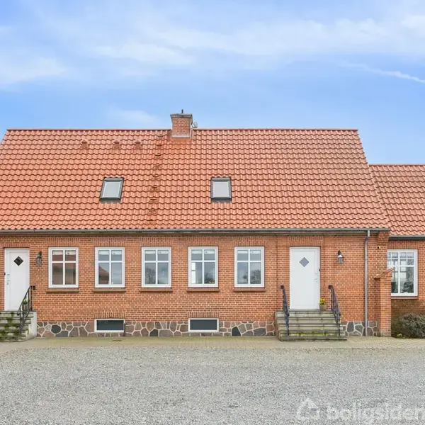 Rødt murstenshus med tegltag og tre kviste. Bygningen har en dør på hver side og fem vinduer i midten. Grusbelagt gårdsplads foran, og træer i baggrunden.