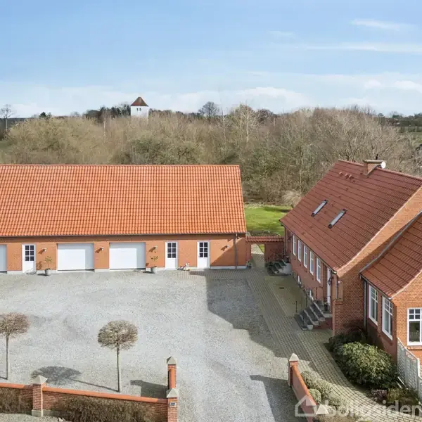 Et rødt murstenshus og garage med orange tegltage står på en grusbelagt gårdsplads. Omgivet af marker og træer, med en hvid kirketårn synligt i baggrunden.