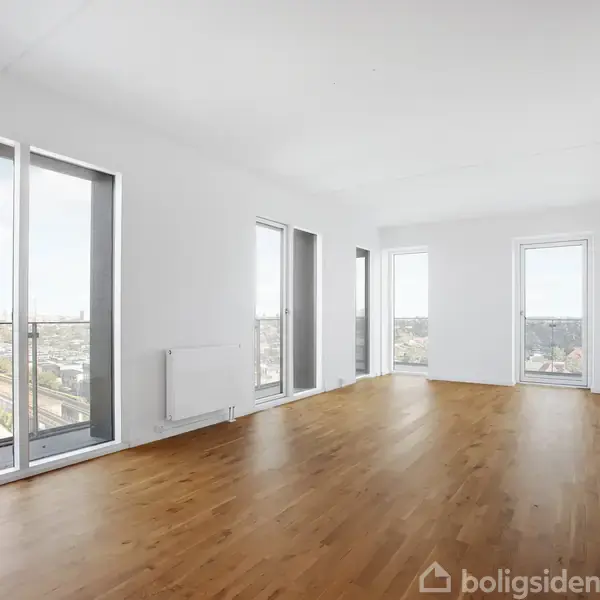 En lys stue med trægulv har store gulv-til-loft vinduer, der giver udsigt over en by. Væggene er hvide, og der er en radiator under vinduerne.