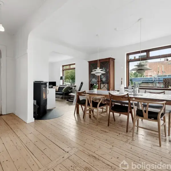 Stort spisebord med stole står på trægulv i spisestue. I baggrunden ses sofaområde. Store vinduer giver udsigt til bygninger udenfor. Lampen hænger over bordet.
