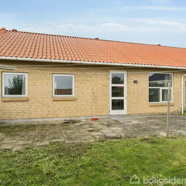 En étplans murstenshus med tre vinduer og en glasdør står på en flisebelagt terrasse. Græsplæner og et tørrestativ omgiver huset under en blå himmel.