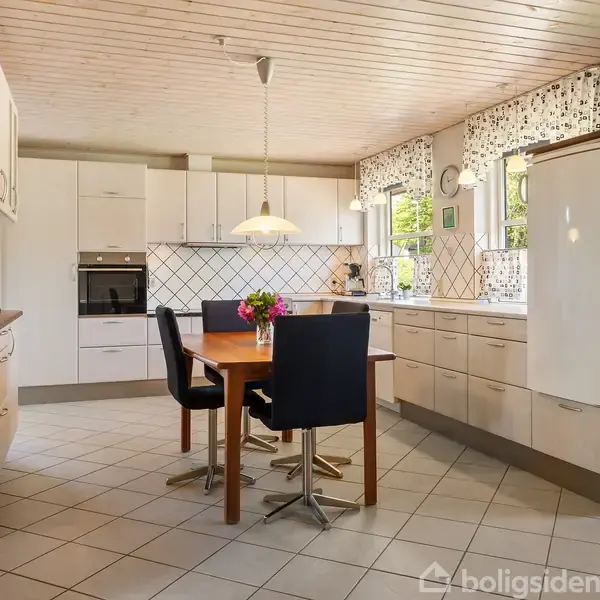 Køkken med hvidt interiør. Spisebord med fire sorte stole står centralt under en hængende lampe. Store vinduer med blomster på bordet og ved siden af køleskabet.