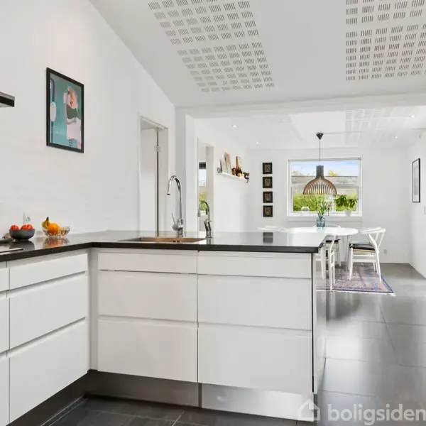 Et moderne køkken i et hvidt rum med sorte gulvfliser og en sort bordplade. I baggrunden er der et spiseområde med et stort vindue og en elegant lampe.