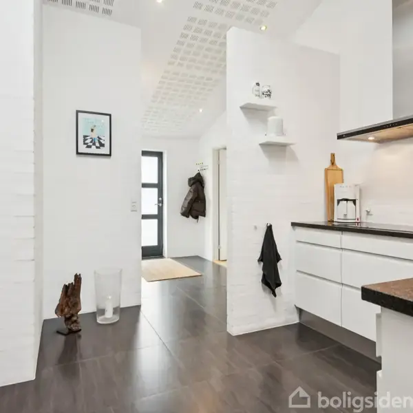 Et moderne køkken- og gangområde med hvide skabe og sort bordplade, udsugningshætte og indbygget ovn. I baggrunden ses en glasdør og væg hylder med dekorationer.