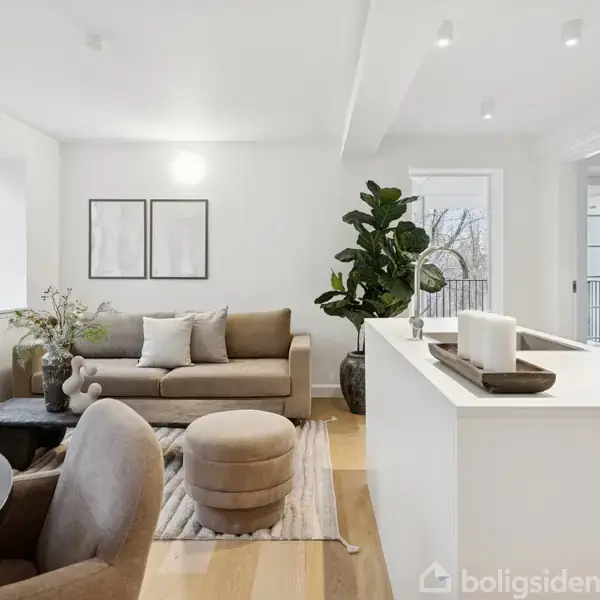 Sofa står i moderne stue ved siden af grøn plante. Skænk med dekorationer og glasdør til balkon i baggrunden. Væggene er hvide, og rummet har trægulv og minimalistisk interiør.