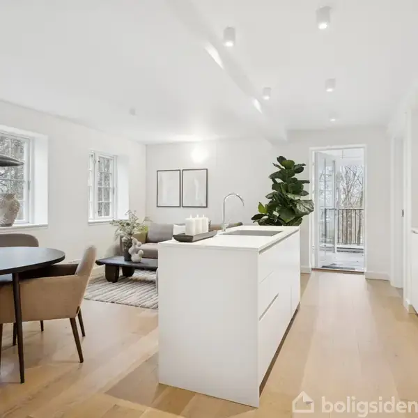 Et moderne køkken-alrum har en stor, hvid køkkenø og spisebord med polstrede stole. En stue med sofa og planter er synlig. Store vinduer giver udsigt til altan og natur.