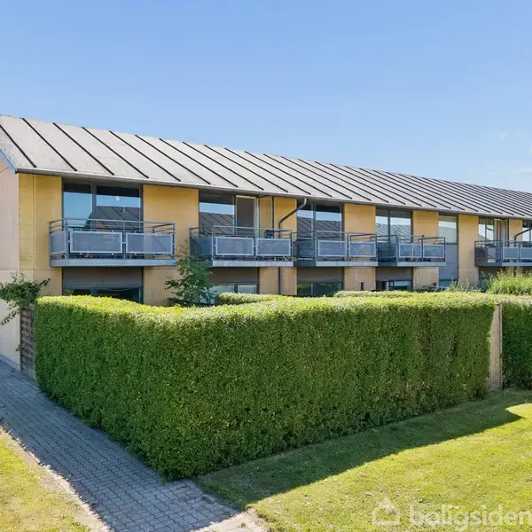 Et moderne rækkehuskompleks står under en blå himmel. Huset har en mørk metaltag og altaner med glasværn. Omgivelserne inkluderer en klippet hæk og velplejet græsplæne.