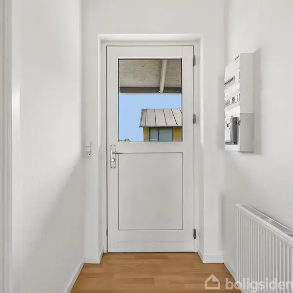 Dør med glasparti fører ud til åben altan med udsigt til et tag. Lys væg, radiator og elpanel i en hvid gang med trægulv. Skarp udendørs belysning.